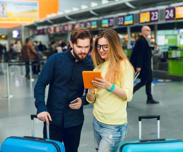 young-couple-is-standing-two-suitcases-airport-she-has-long-hair-glasses-sweater-jeans-he-wears-beard-black-shirt-with-pants-they-are-reading-tablet(1)